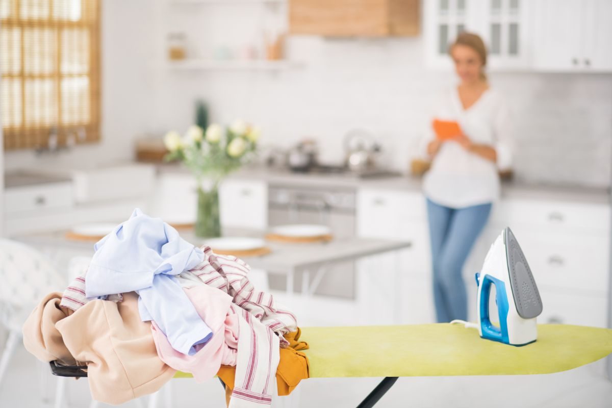 asse da stiro con vestiti in primo piano e sullo sfondo una donna con smartphone